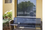 Metal bench on a covered exterior porch in front of a window with vertical blinds and surrounding landscaping.