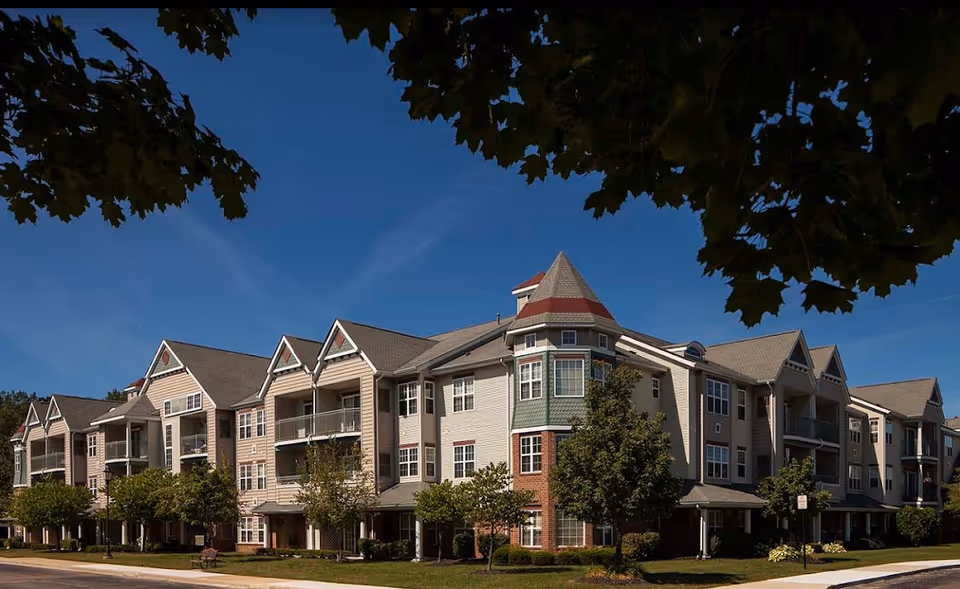 Exterior front view of a multi-story senior living building with balconies, gables, and landscaped grounds under a blue sky.