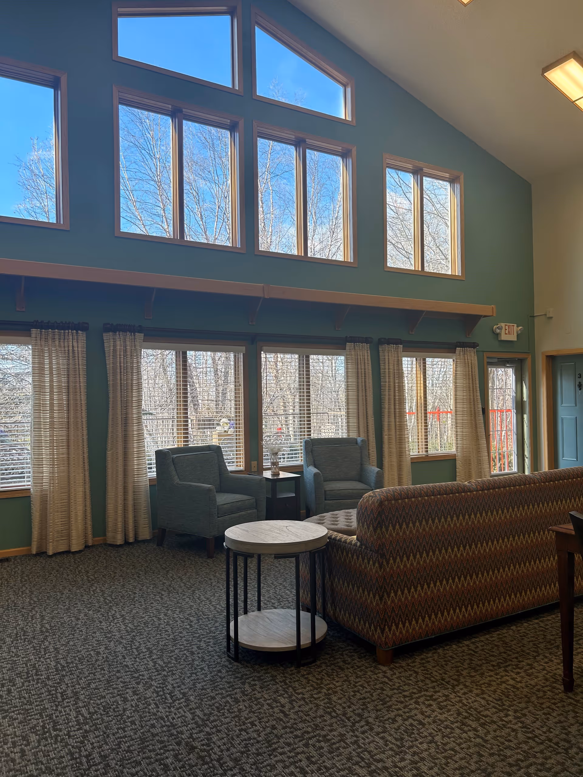 A spacious living room area with large windows letting in natural light. The room features a patterned sofa, two blue armchairs, a round side table, and curtains on the windows. The walls are painted green, and the ceiling is vaulted with additional upper windows. Outside, leafless trees are visible through the windows.