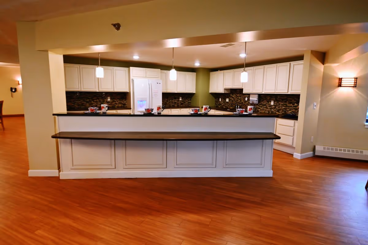 Open communal kitchen with a long breakfast bar, white cabinets, pendant lights, and wood floors.