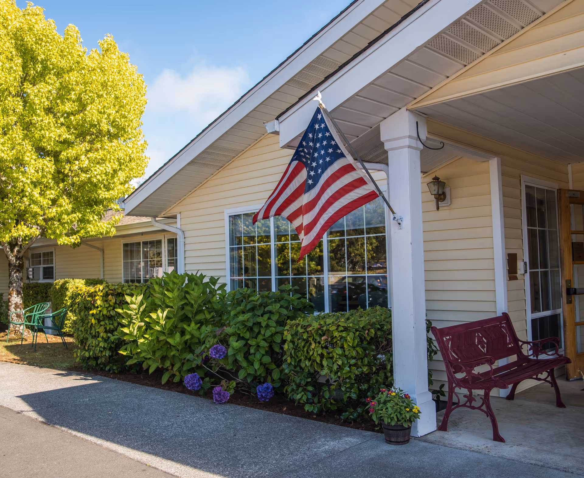 Exterior view of a senior living facility with beige siding, a large window, and an American flag mounted on a white column. There is a red bench on the porch, a potted plant, green bushes with purple flowers, and a tree with bright green leaves under a clear blue sky.