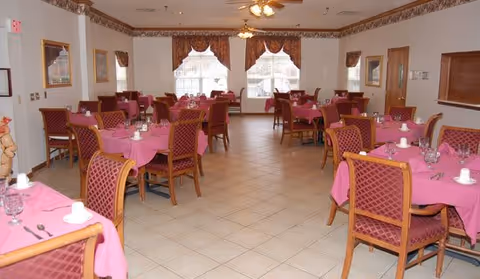 A dining room with multiple tables covered in pink tablecloths, each set with white cups, glasses, and silverware. The room has tiled floors, large windows with valances, and wooden chairs with patterned upholstery. Ceiling fans and framed pictures are visible on the walls.