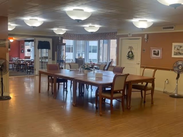 A spacious dining room with wooden floors and a large rectangular wooden table surrounded by chairs. The room is well-lit with ceiling lights and has windows with valances letting in natural light. There are fans on stands placed around the room, and a wreath hangs on a door in the background. Another smaller dining area is visible through an open doorway.