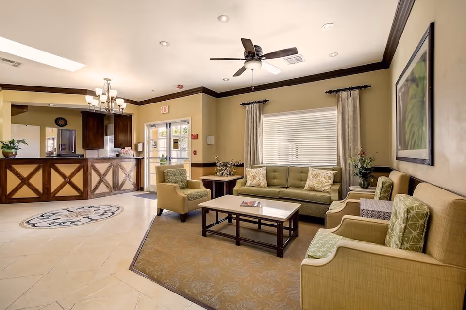 A well-lit senior living facility common area with beige walls and tiled floors. The room features a green sofa with floral pillows, two green armchairs with patterned cushions, a wooden coffee table, and a side table with a flower vase. A ceiling fan and chandelier provide lighting. In the background, there is a reception desk with wooden paneling and a glass door entrance.