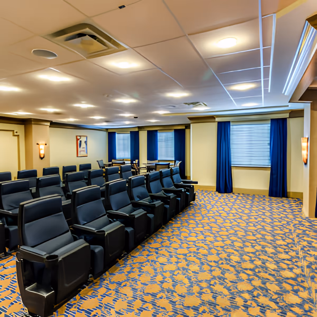Interior view of a theater-style room with multiple rows of black reclining chairs arranged on a patterned carpet. The room has beige walls with blue curtains covering the windows and soft ceiling lighting.
