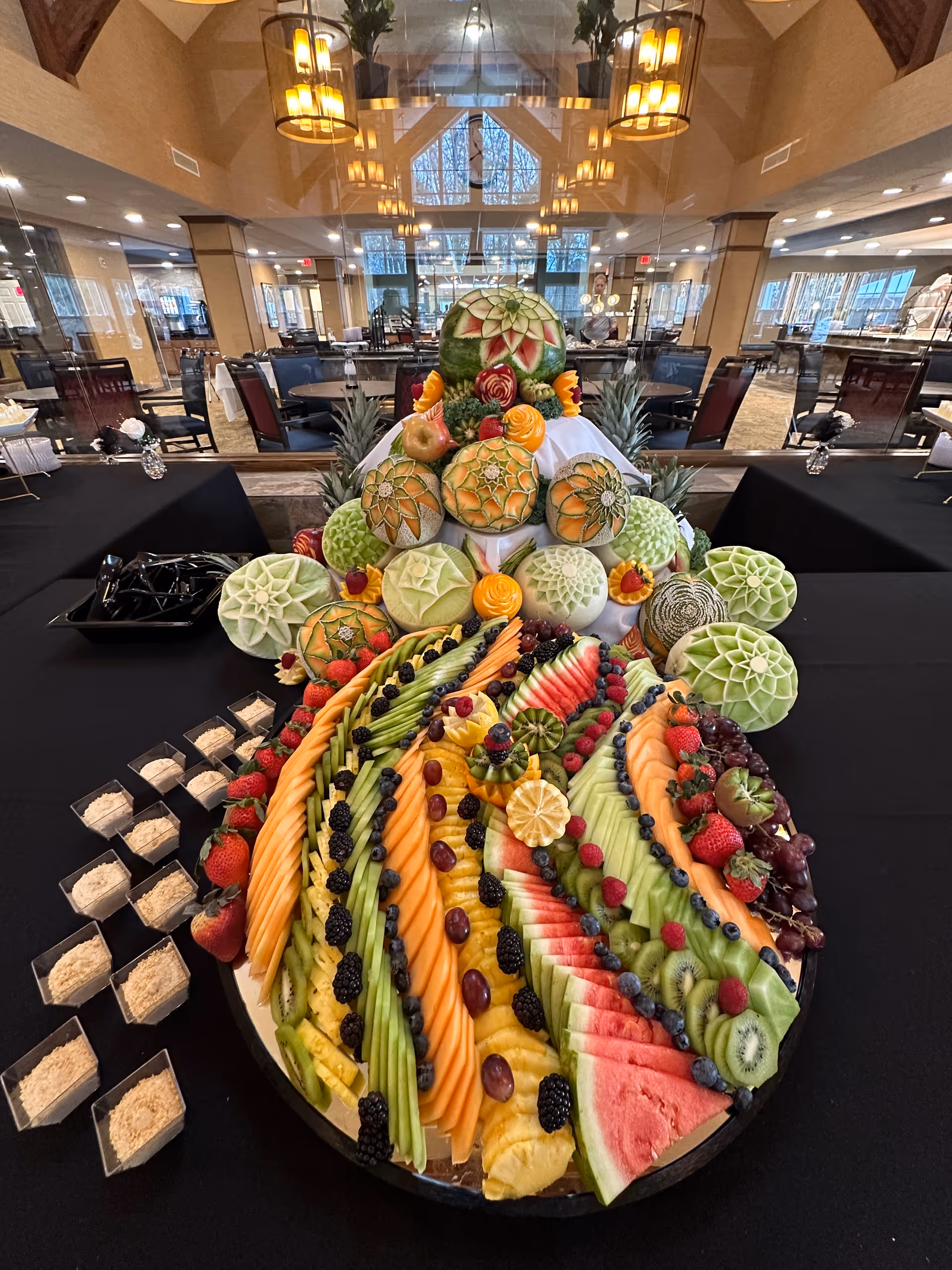 A large, colorful fruit display arranged on a table inside a dining area. The display features intricately carved melons stacked in a pyramid shape at the back, surrounded by an assortment of sliced fruits including watermelon, cantaloupe, pineapple, kiwi, grapes, strawberries, blackberries, blueberries, and raspberries. The dining area has tables and chairs, large windows, and warm lighting fixtures hanging from the ceiling.