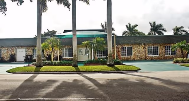 Single-story assisted living building front with a stone facade, palm trees, and a green awning over the entrance.