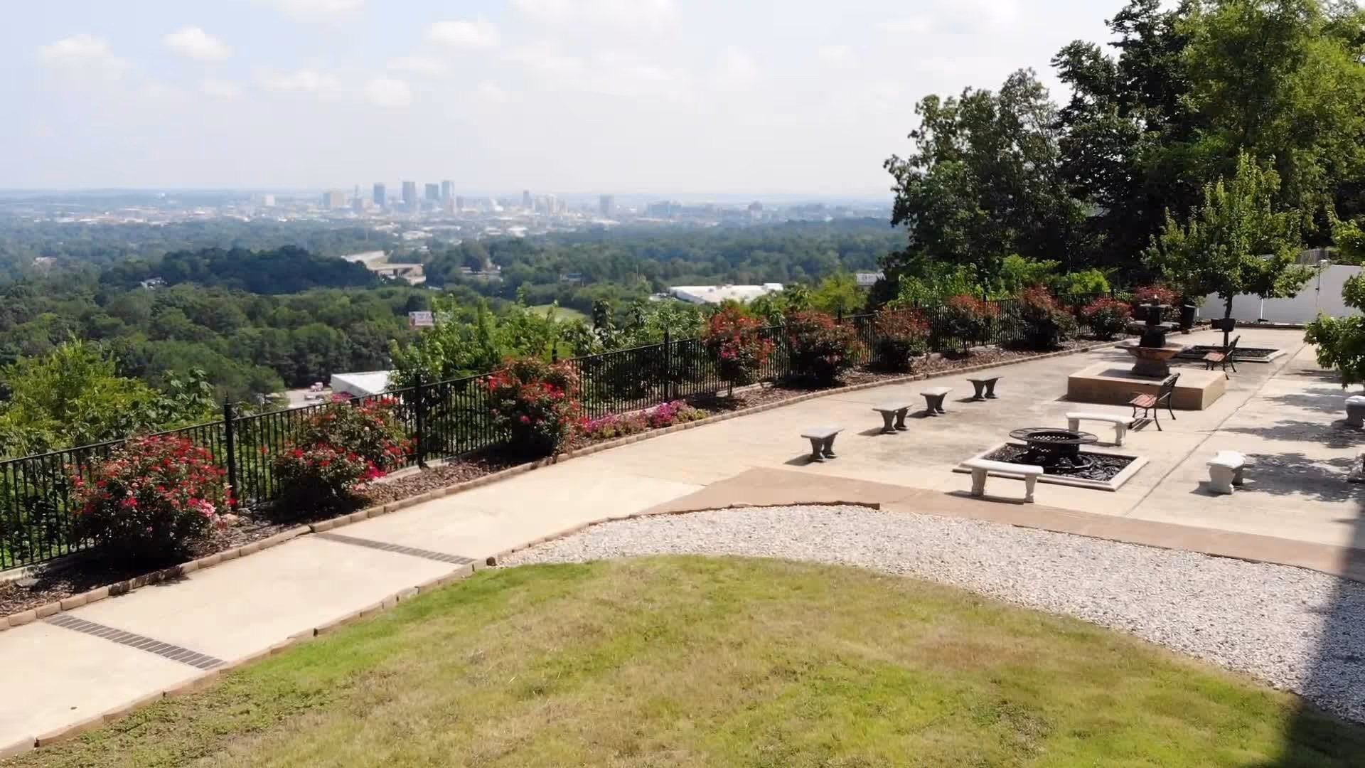 Outdoor patio area with benches, a fire pit, and a fountain surrounded by trees and flowering bushes, overlooking a distant city skyline under a partly cloudy sky.