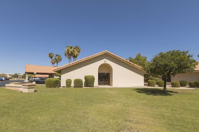 Exterior view of a single-story building with a beige stucco finish and a red tile roof, surrounded by green grass, trimmed bushes, a tree, and palm trees in the background under a clear blue sky.