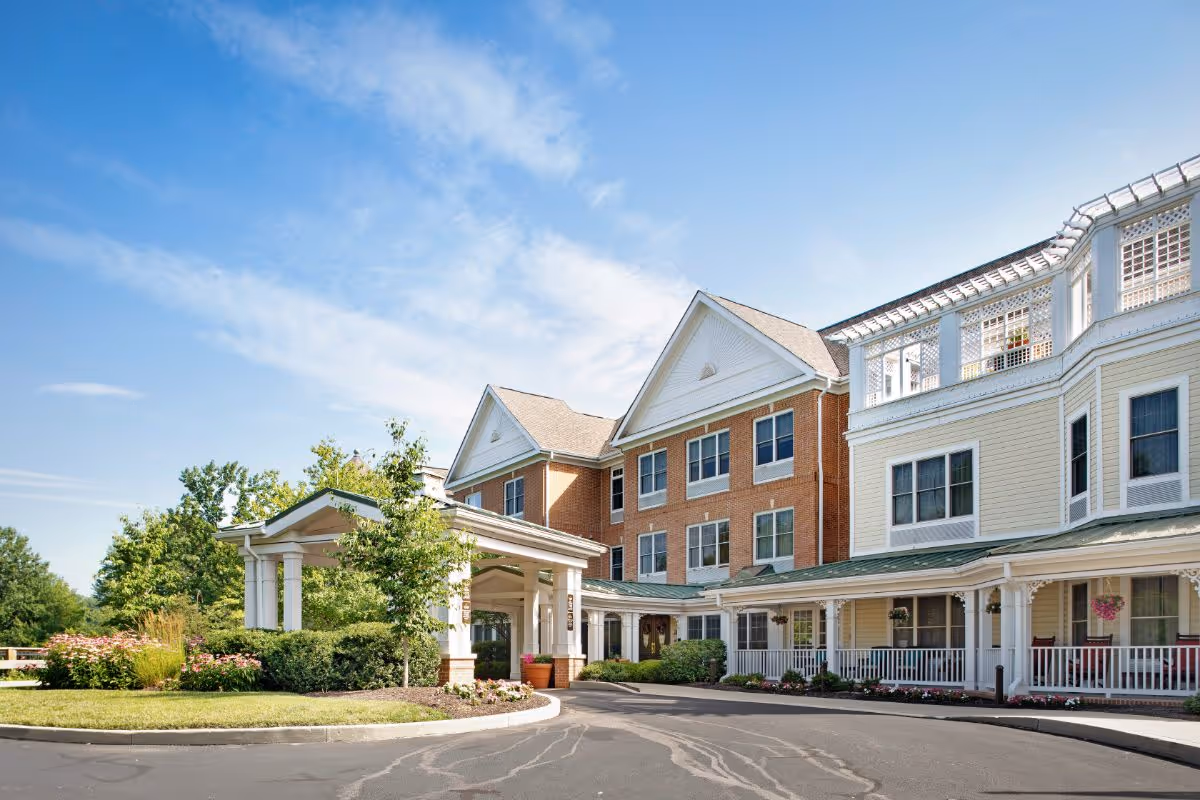 Exterior view of a senior living facility building with a covered entrance, landscaped greenery, and a clear blue sky.