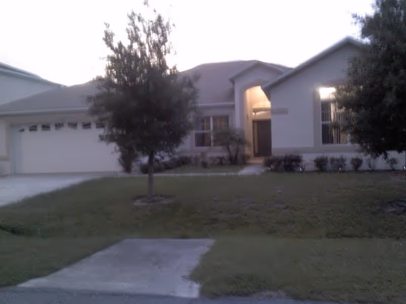 Front exterior view of a single-story residential-style building with a garage, a front door with a small porch, two trees in the front yard, and a lawn area.