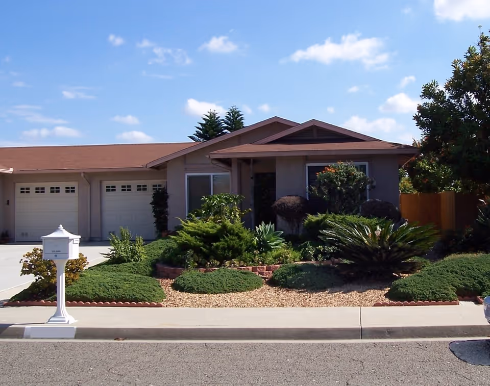 Single-story suburban house front with a three-car garage, landscaped yard and white mailbox under a blue sky.