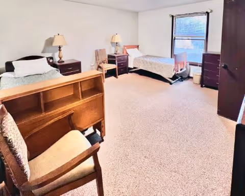 A clean and simple bedroom with two single beds, each with a bedside table and lamp. There is a wooden chair in the foreground and a window with blinds on the far wall. The room has beige carpeting and light-colored walls.