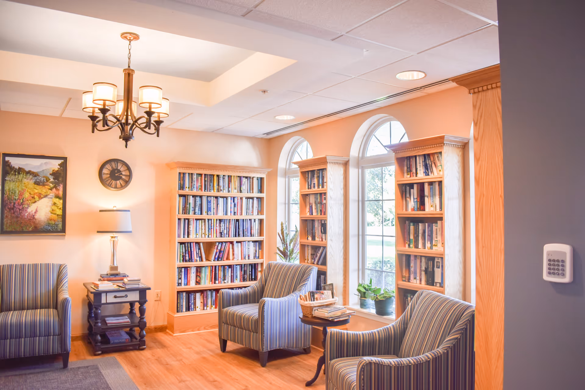 A cozy reading area with three striped armchairs, wooden bookshelves filled with books, large arched windows letting in natural light, a small side table with a basket and books, a table lamp on a small dark wooden side table, a wall clock, and a landscape painting on the wall.