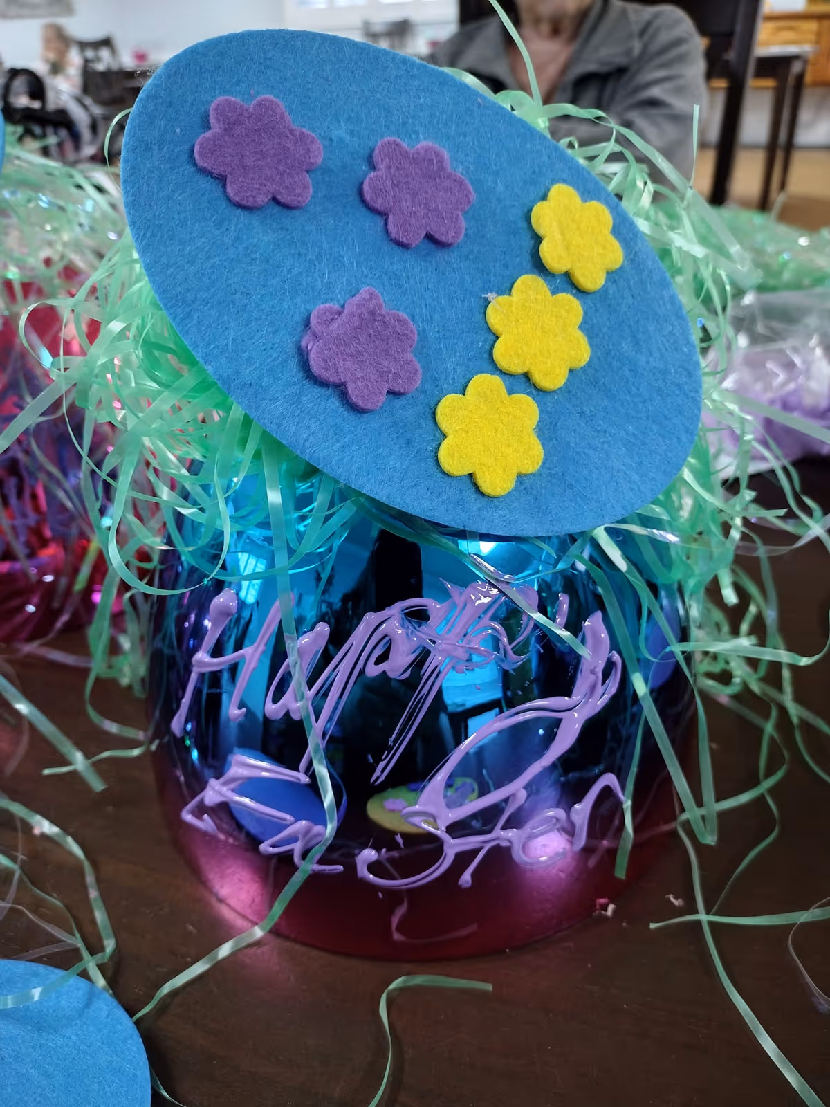 Close-up of a colorful decorated centerpiece jar topped with a blue felt circle adorned with purple and yellow flower shapes and green shredded paper.