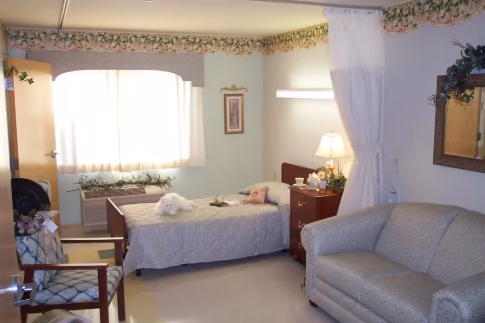 A cozy senior living bedroom with a single bed, nightstand and lamp, a loveseat and chair near a curtained window.