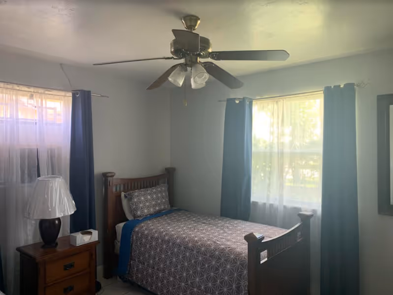 A small bedroom with a single wooden bed covered with a patterned gray and blue bedspread. There is a wooden nightstand next to the bed with a lamp and a tissue box on it. The room has two windows with sheer white curtains and dark blue drapes. A ceiling fan with lights is mounted on the ceiling.