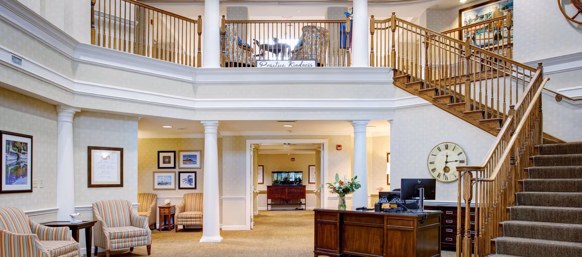 Spacious two-story reception lobby with seating areas, white columns, a wooden staircase, and a front desk.