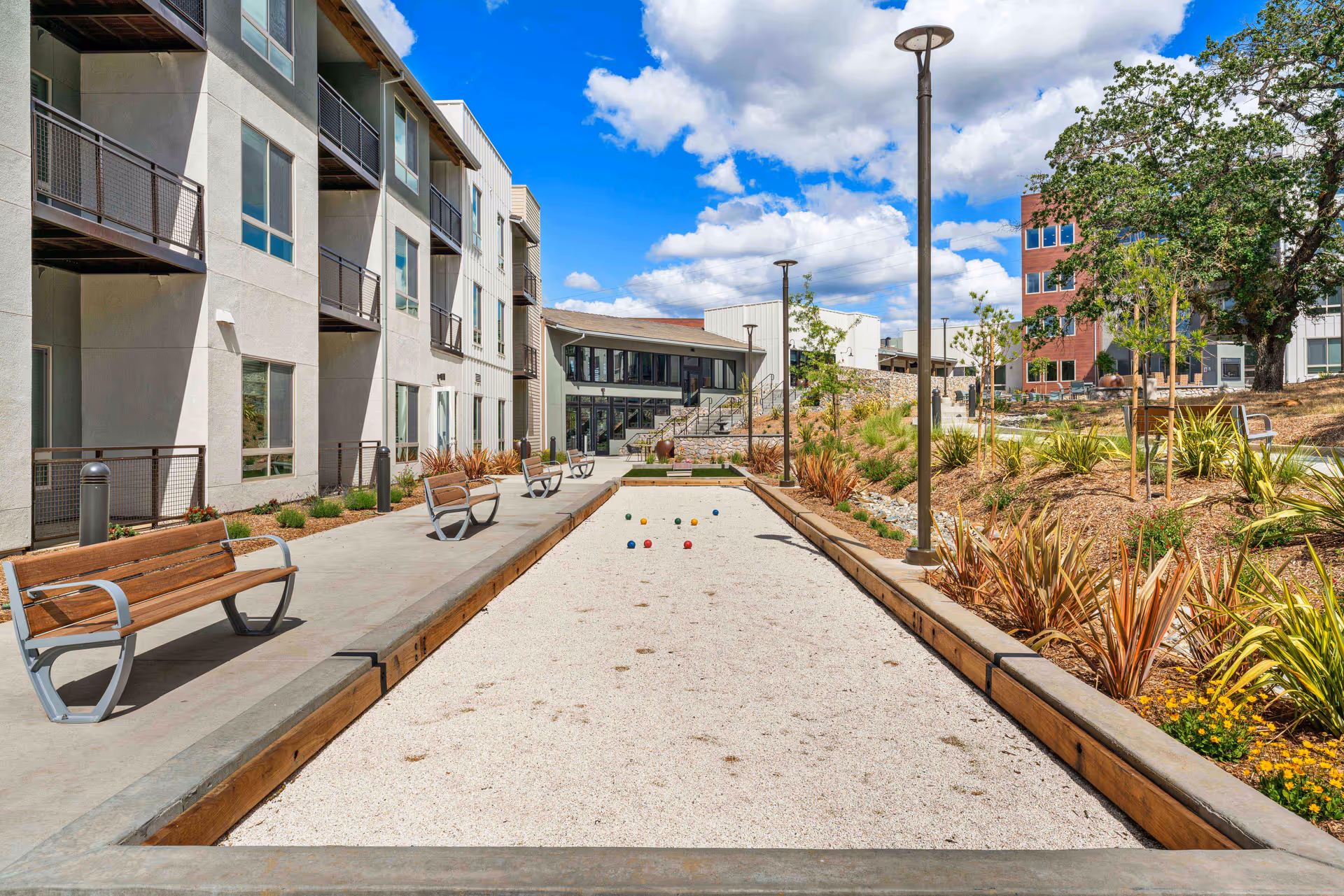 Outdoor bocce ball court with benches along the side, surrounded by landscaped plants and trees. Modern multi-story buildings are visible on the left and in the background under a partly cloudy blue sky.