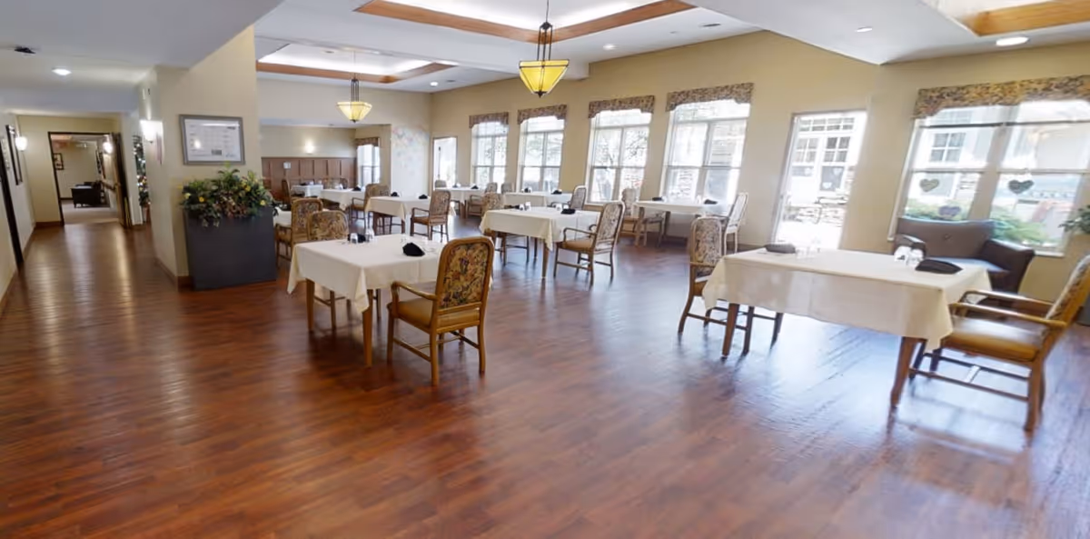 A spacious dining room with wooden floors, multiple tables covered with white tablecloths, and chairs with floral upholstery. Large windows on one side let in natural light, and there are ceiling lights and wall sconces providing additional lighting. A hallway is visible on the left side of the room.