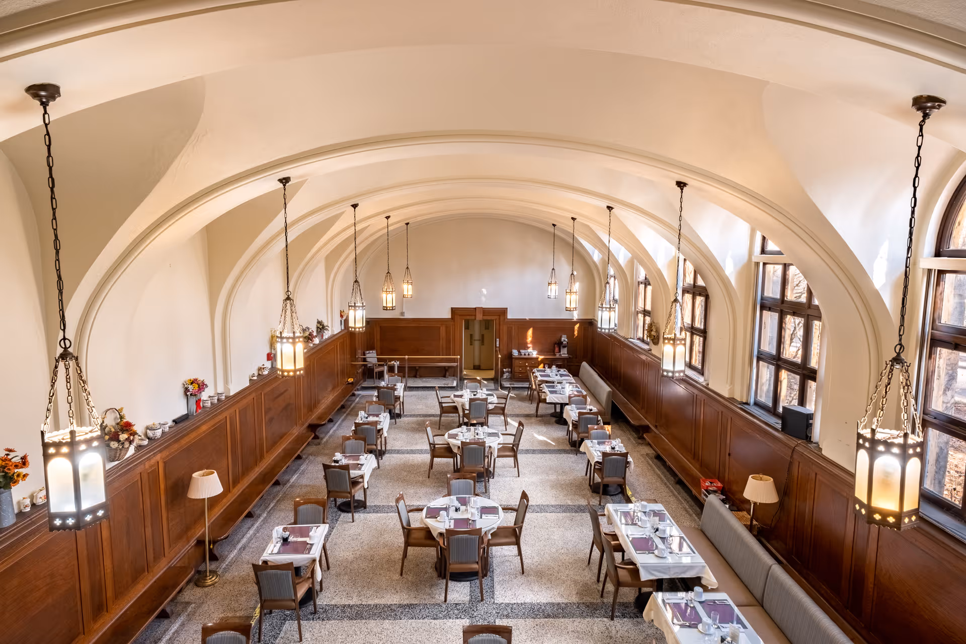 A spacious dining room with arched ceilings and large windows letting in natural light. The room features multiple tables set with placemats, napkins, and silverware, surrounded by chairs. Pendant lights hang from the ceiling, and there are wooden panels along the walls with decorative items and lamps.