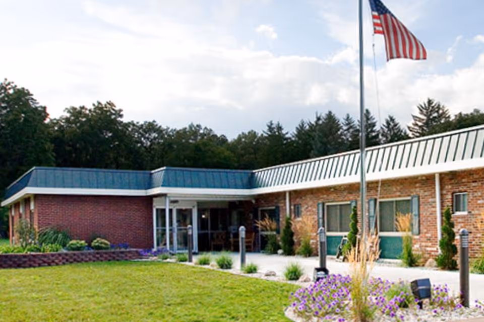 Single-story brick senior living facility front entrance with an American flag and landscaped lawn.
