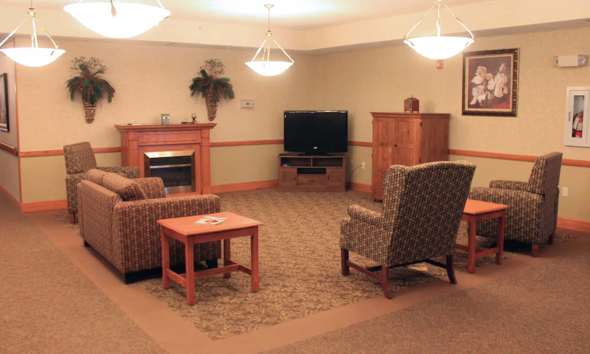 A cozy living room area with patterned armchairs and a sofa arranged around a small wooden table. The room features a wooden fireplace with decorative plants above it, a flat-screen TV on a wooden stand, a wooden cabinet, and a framed floral painting on the wall. The lighting is provided by ceiling-mounted pendant lights.