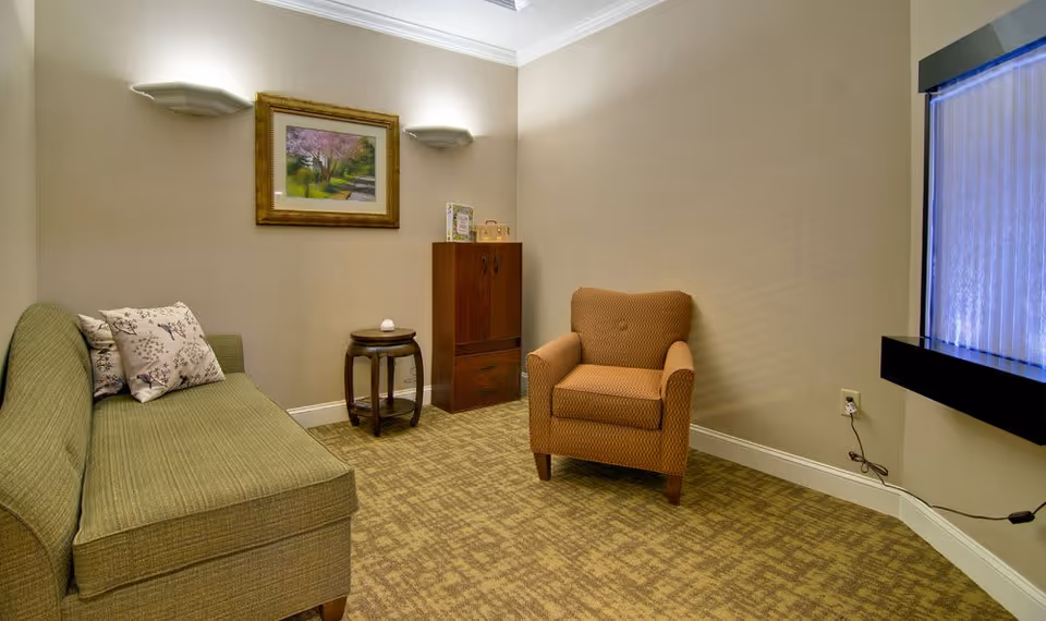 Carpeted sitting room with a green sofa, an upholstered armchair, a round side table and cabinet beneath framed artwork and a vertical illuminated water feature on the right wall.