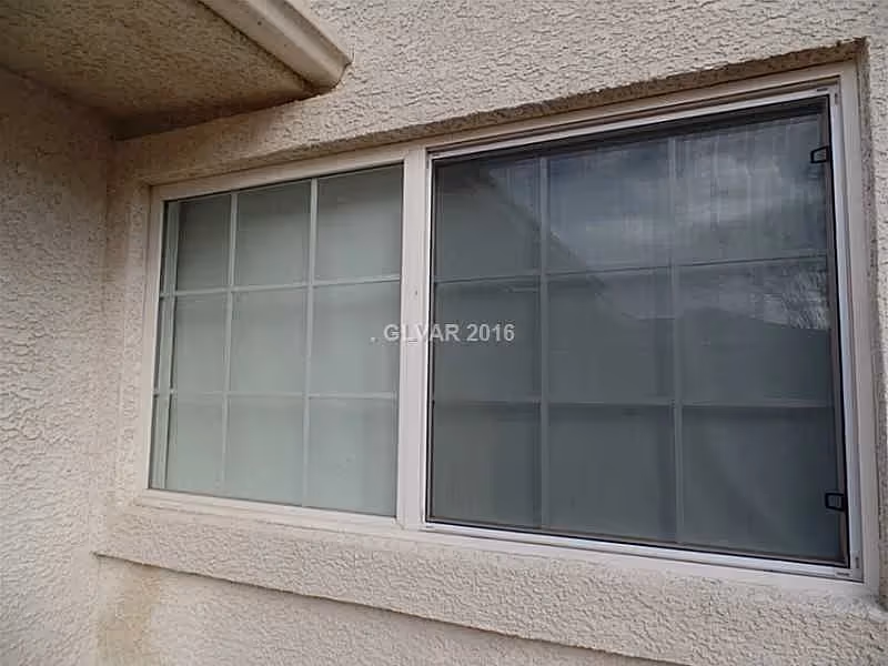 A close-up view of a double-pane window with white frames set in a textured beige exterior wall. The window has grid patterns dividing the glass into smaller sections. The left pane is frosted or covered, while the right pane is clear with a reflection of the sky and nearby structures.