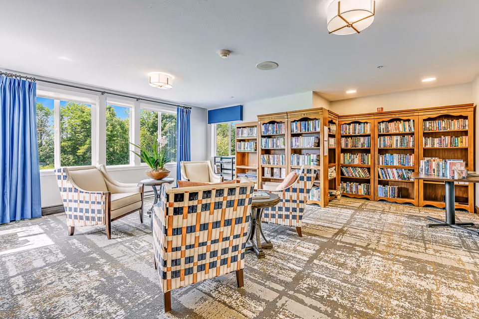 A bright and spacious library or reading room with large windows letting in natural light. The room features several wooden bookshelves filled with books, four cushioned armchairs with a checkered pattern arranged around a small round table, and blue curtains framing the windows. The carpet has a modern pattern, and there are ceiling lights providing additional illumination.
