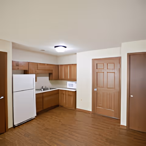 A kitchen area with wooden cabinets, a white refrigerator, a microwave on the countertop, and a sink. The floor is wooden, and there are two closed wooden doors visible on the right side of the image. The walls are painted white, and a ceiling light fixture is centered above.