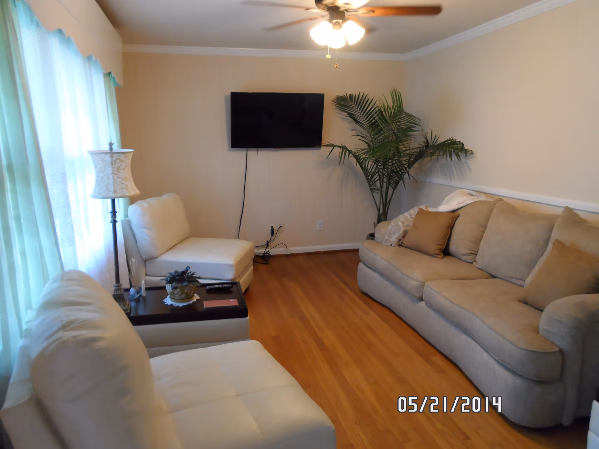 Cozy living room with a beige sofa, white chairs, wall-mounted TV, potted palm, floor lamp, and hardwood floors.