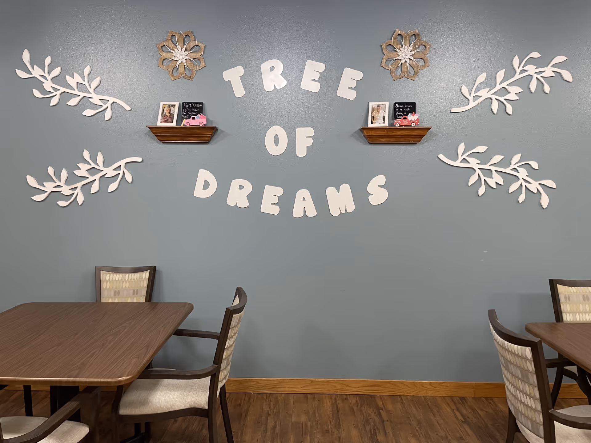 Interior wall decorated with the phrase 'TREE OF DREAMS' in large white letters. Two small wooden shelves hold framed photos and small pink toy cars. White decorative floral and leaf designs are arranged around the text on a blue-gray wall. Below are wooden tables and cushioned chairs with patterned upholstery on a wooden floor.