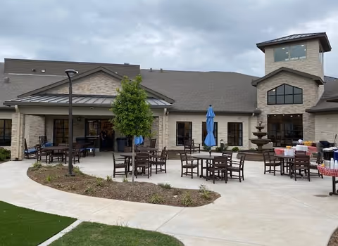 Outdoor patio area of a senior living facility with multiple wooden tables and chairs arranged on a concrete surface. There are blue umbrellas over some tables, a small tree planted in a landscaped area, and a multi-tiered water fountain near the building. The building has light-colored brick walls, large windows, and a tower-like structure on the right side under a cloudy sky.