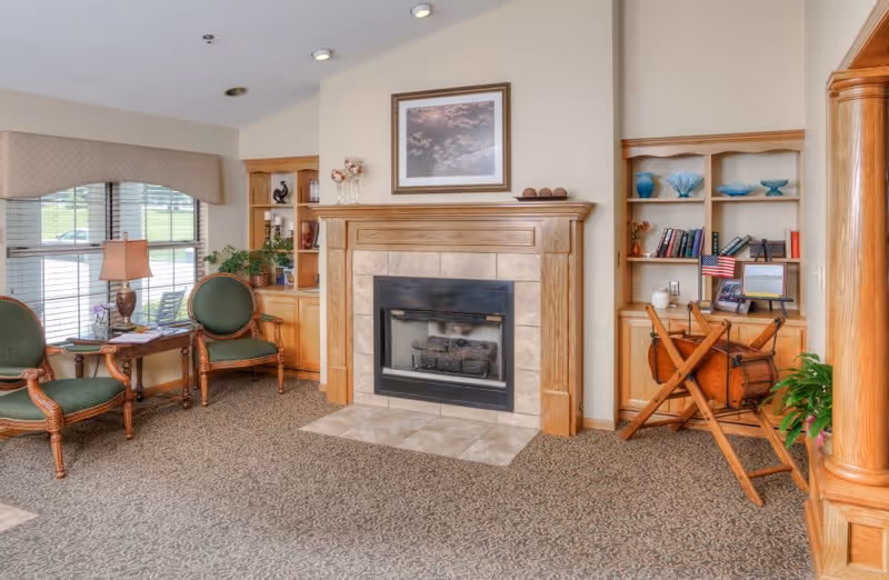 A cozy living room area with a fireplace in the center, flanked by built-in wooden shelves filled with books and decorative items. Two green upholstered chairs with wooden frames are placed near a window with blinds and a valance. A small wooden table with a lamp and a plant sits between the chairs. The carpet has a patterned design, and the walls are painted a light color. There is a framed picture hanging above the fireplace.
