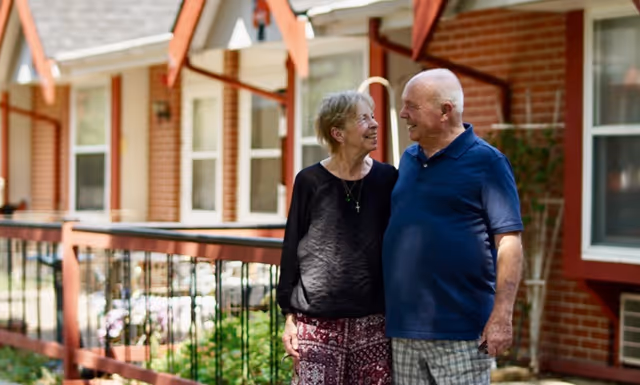 An elderly couple standing outside in front of a brick building with white-framed windows and red trim. They are smiling at each other and appear happy, with a railing and greenery visible in the background.