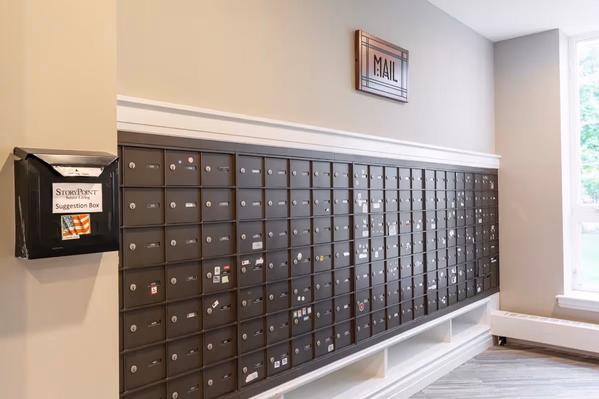 A wall of multiple black mailboxes with small locks and labels, mounted in a senior living facility. To the left, there is a black suggestion box labeled 'StoryPoint Senior Living Suggestion Box' with an American flag sticker. Above the mailboxes, a decorative sign reads 'MAIL'. A large window on the right side lets in natural light.