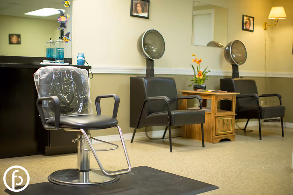 Interior of a salon area with a black salon chair covered with a plastic cape in the foreground, two black chairs with hair dryers behind them, a wooden side table with a flower arrangement, and framed pictures on the beige walls.