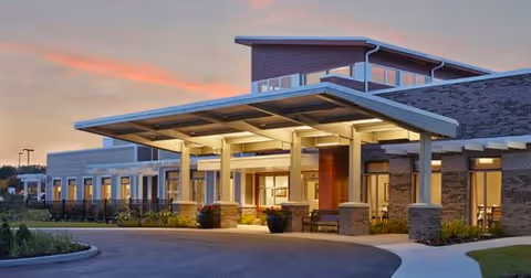 Exterior view of The Springs at Lafayette facility at dusk, showing the entrance with a covered drop-off area, illuminated by warm lights. The building has a modern design with large windows and a combination of brick and siding materials.