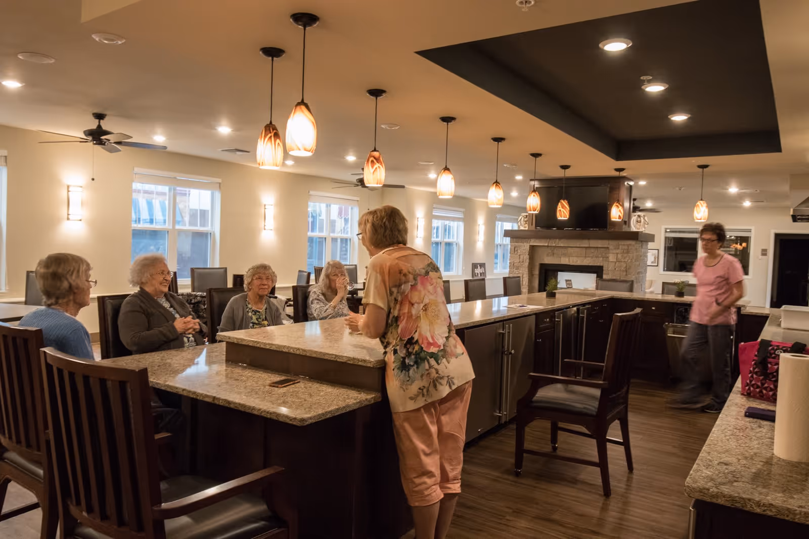 A group of elderly women sitting and standing around a large kitchen island with granite countertops in a well-lit assisted living facility. The room features pendant lights, ceiling fans, large windows, and a stone fireplace in the background. One woman is standing and talking to the seated group, while another woman walks in the background.