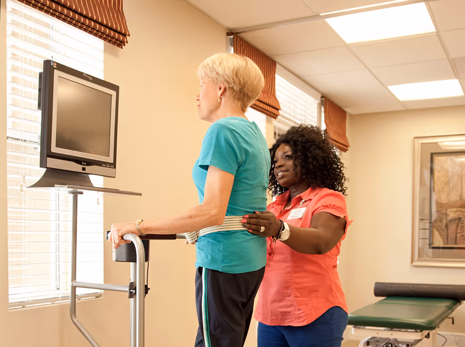 A healthcare worker assists an elderly woman using a walker in a rehabilitation room with a monitor in front of them and windows with blinds and brown valances in the background.