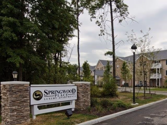 Entrance sign for Springwood Place Luxury Apartments with stone pillars on either side, surrounded by trees and landscaped greenery, with apartment buildings visible in the background under a cloudy sky.