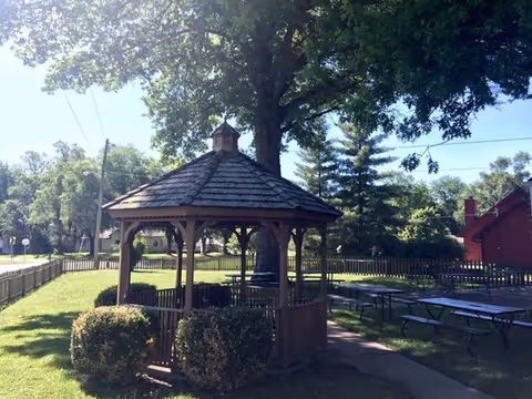Outdoor area with a wooden gazebo surrounded by trimmed bushes, picnic tables on a grassy lawn, large trees providing shade, and a wooden fence enclosing the space under a clear blue sky.