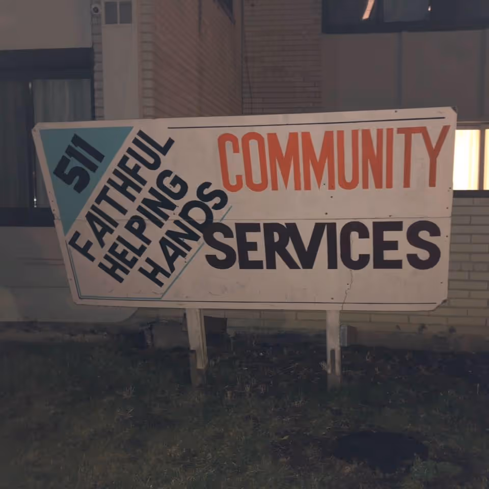 A large outdoor sign for Faithful Helping Hands Community Services, positioned on a grassy area in front of a brick building at night.