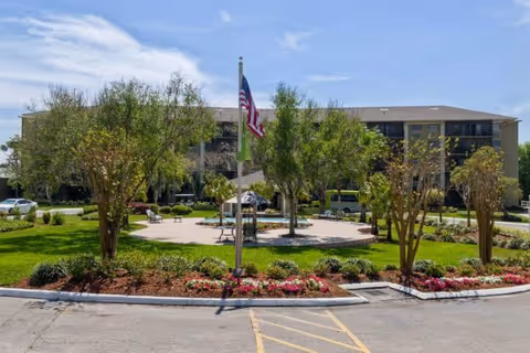Front exterior of a multi-story senior living building with a circular landscaped entrance, flagpole, and benches.