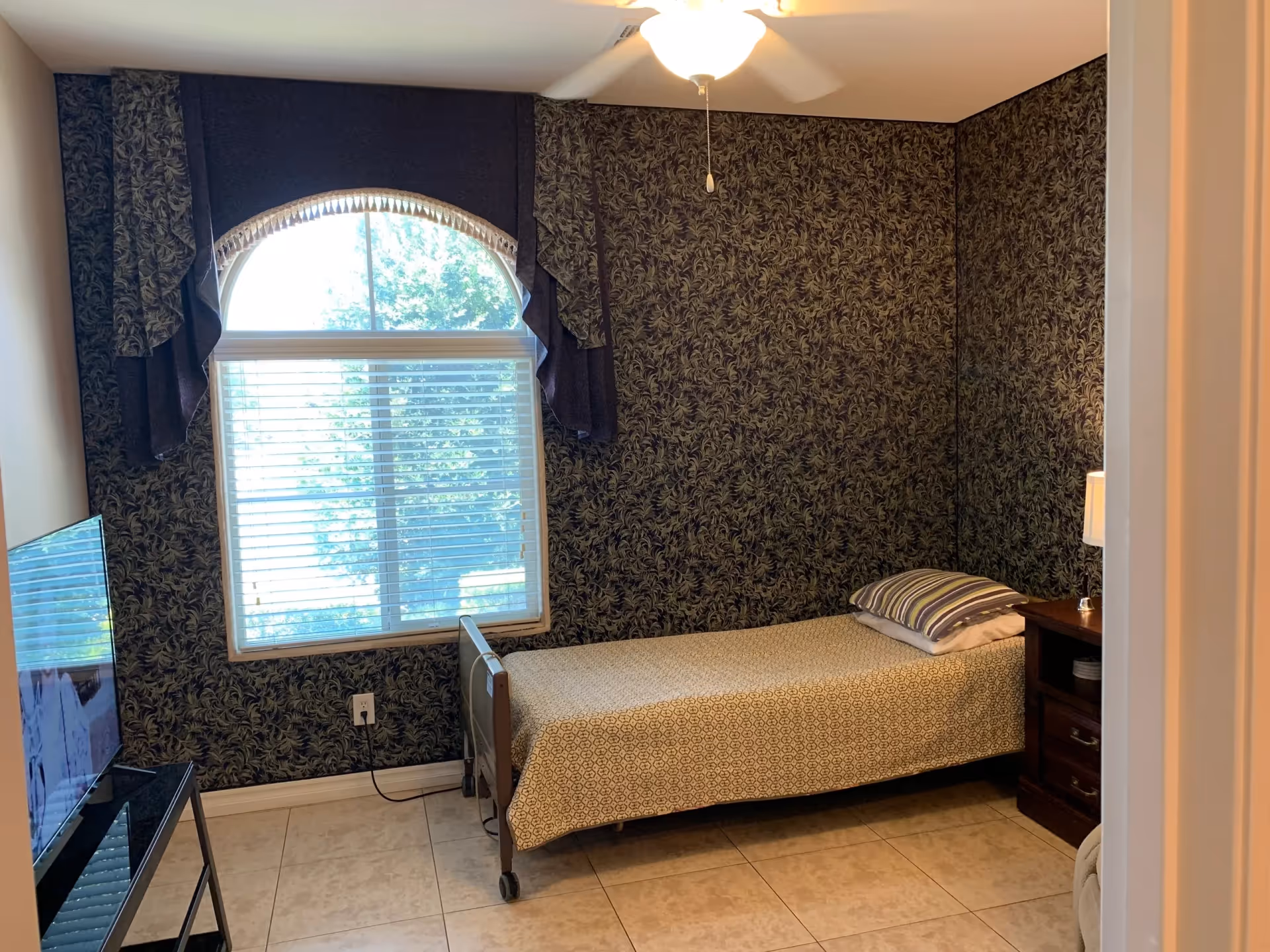A single bedroom in a senior living facility with patterned wallpaper, window, bed, nightstand, and TV.
