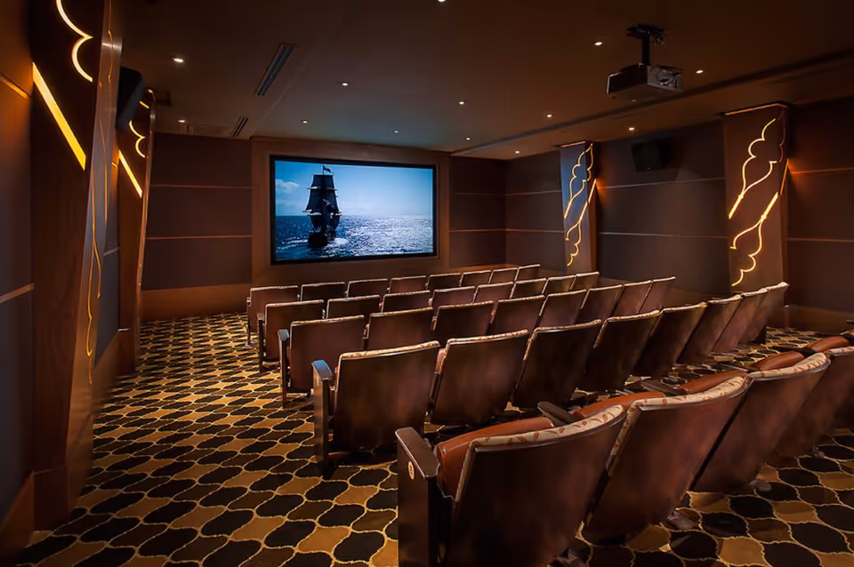 A small private movie theater with rows of upholstered seats facing a large screen showing a sailing ship.