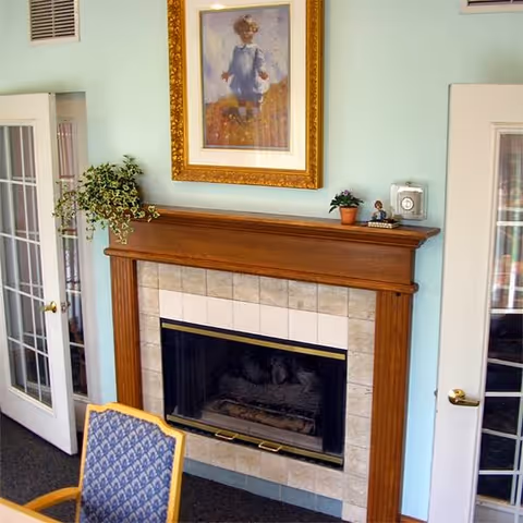 Interior room showing a wood-framed tile fireplace with a framed painting above, plants and decor on the mantel, glass-paned French doors, and a patterned chair in the foreground.