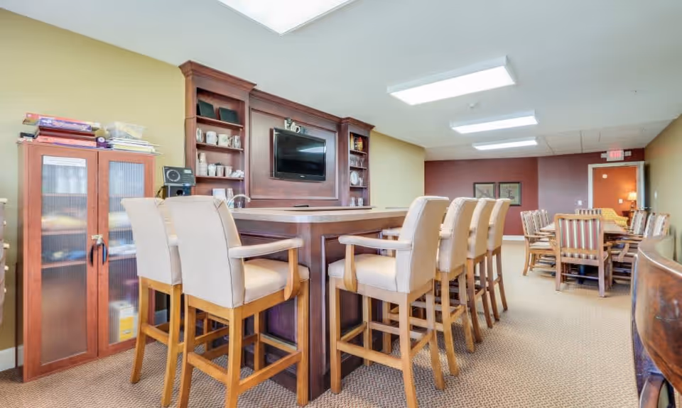 A spacious common area with a large wooden table surrounded by cushioned high chairs. The room features a built-in wooden shelving unit with a mounted flat-screen TV and various decorative items. In the background, there is another dining table with wooden chairs, and the walls are painted in warm tones of yellow and maroon. The ceiling has recessed lighting panels.
