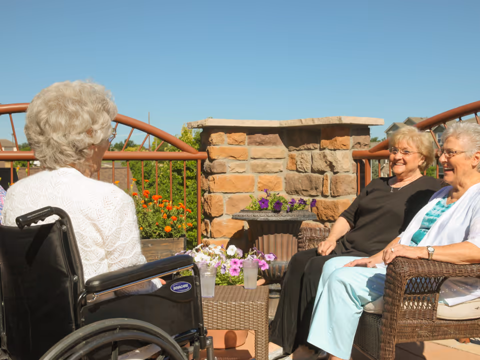 Three elderly women sitting and chatting outdoors on a sunny day. One woman is in a wheelchair wearing a white sweater, and the other two women are seated on wicker chairs with cushions. There are flowers and a stone wall in the background under a clear blue sky.